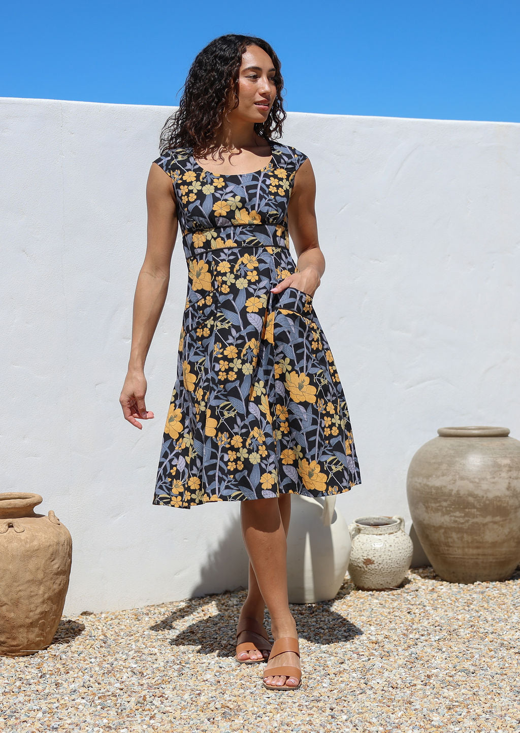Model stands with hand in pocket wearing a retro floral cotton dress with scooped round neckline, cap sleeves and piped details at the waist and pockets