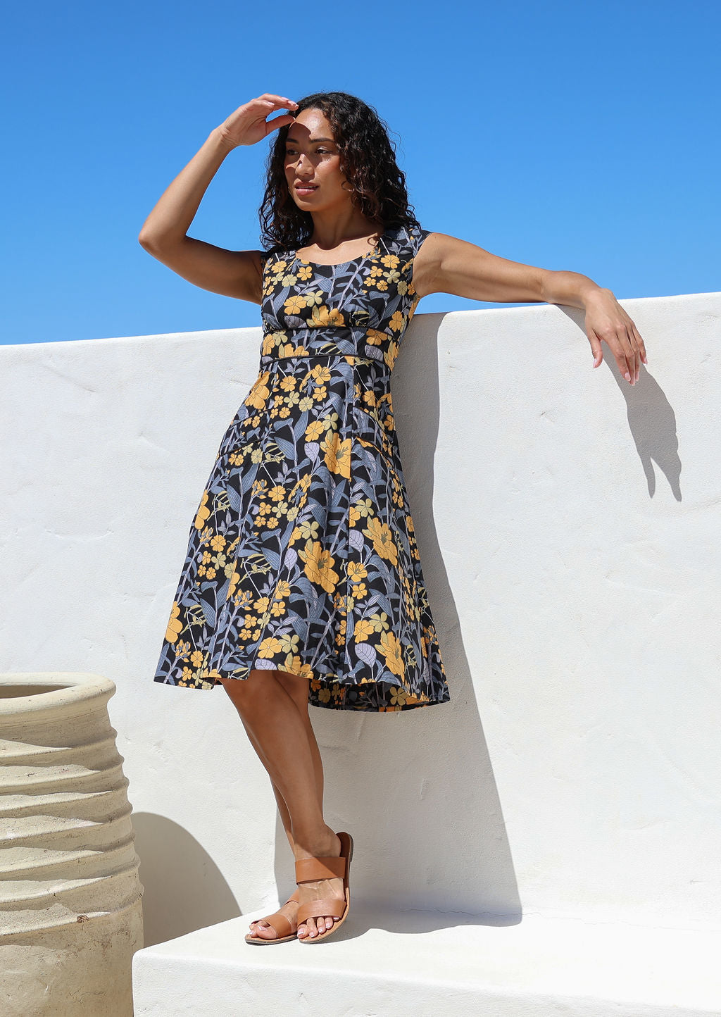 Model stands in sun wearing a 100% cotton knee length dress with scooped round neckline, cap sleeves and piping at the waist and pocket