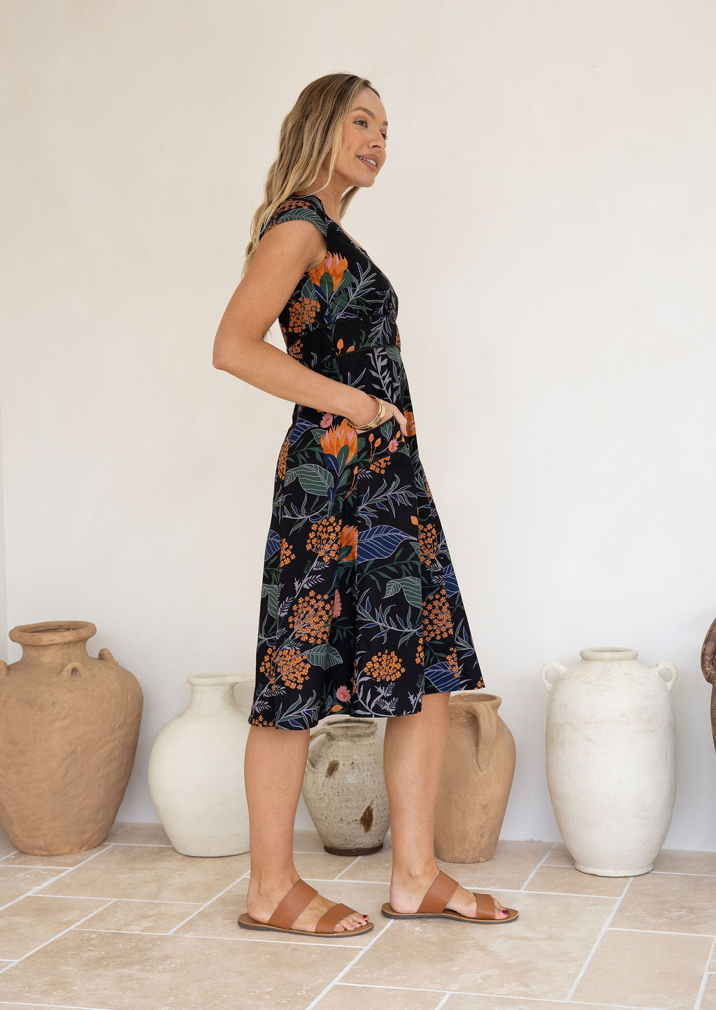 Woman wearing a floral cotton dress with cap sleeves, piped details and hands in pocket, standing in front of ceramic vases.