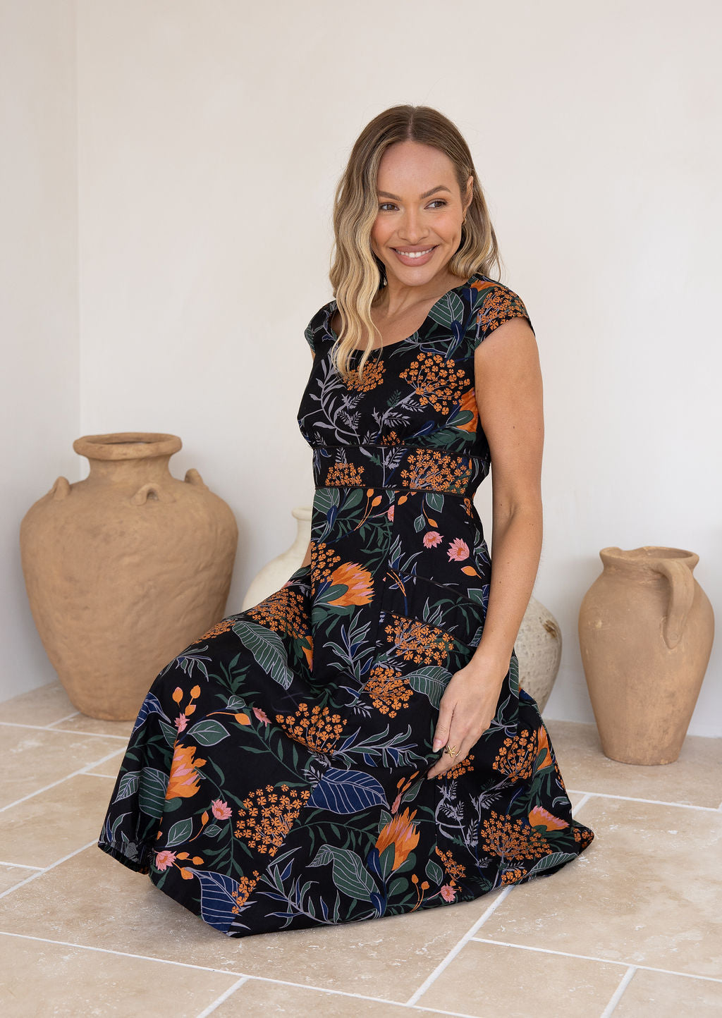 Model sitting in a retro inspired knee length cotton dress with piped detailing in black with orange and pink florals.