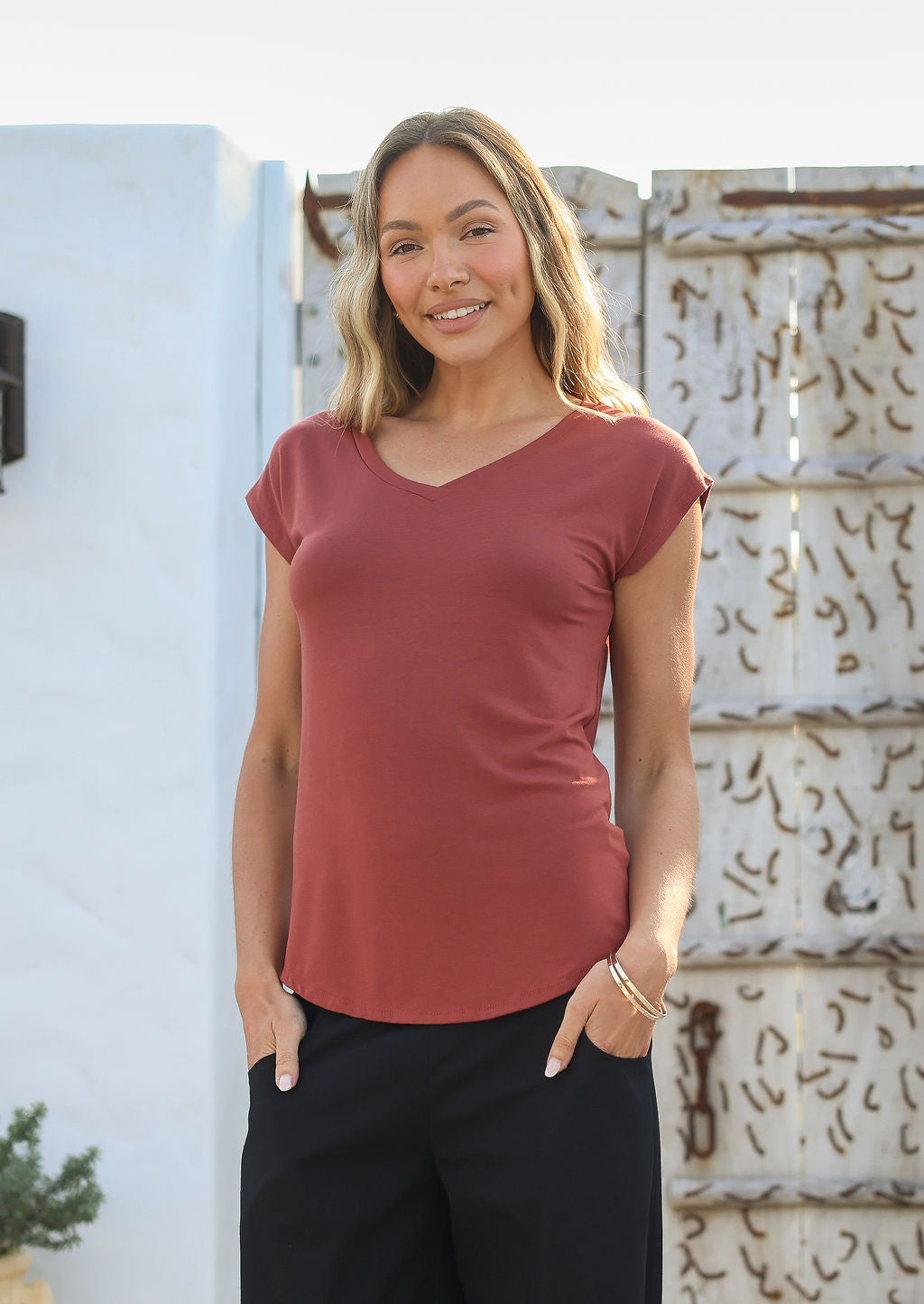 Woman wearing a 100% rayon terracotta V-neck T-shirt and black pants, standing against a wall.