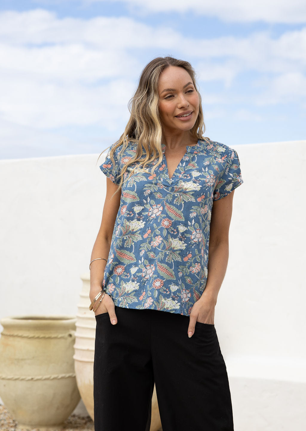 Woman stands with hands in pocket wearing a sweet floral blouse featuring ruched shoulders and V neckline.