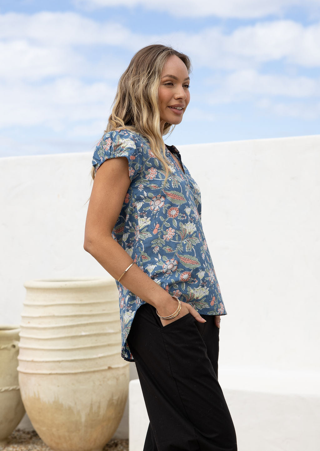 Woman stands sideways in loose fit, blue floral cotton top with elastic trimmed short sleeves and a longer back hem.