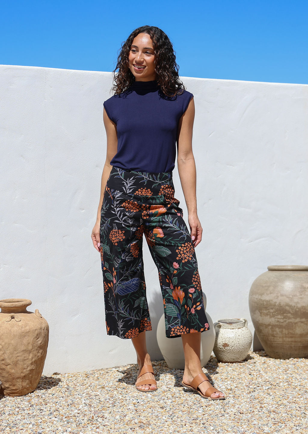 Model standing in sun wearing a 100% cotton capri style wide leg pants in black with bold botanical print and side pockets