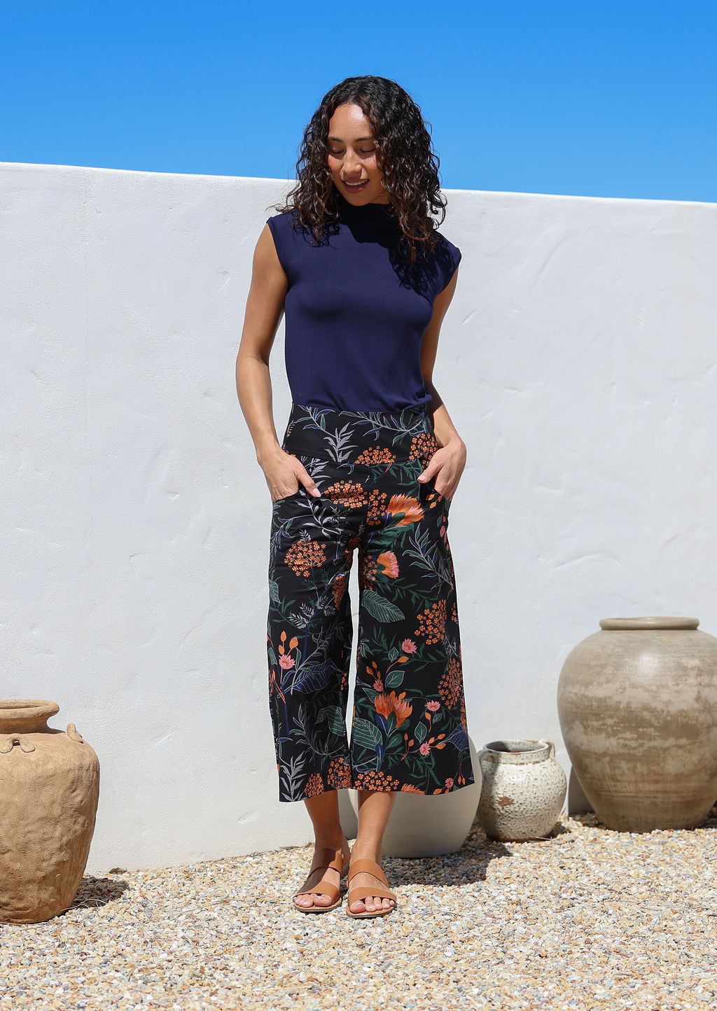 Woman standing with hands in pockets wearing a high waisted wide leg capri style pants featuring beautiful botanical print with pink and orange florals