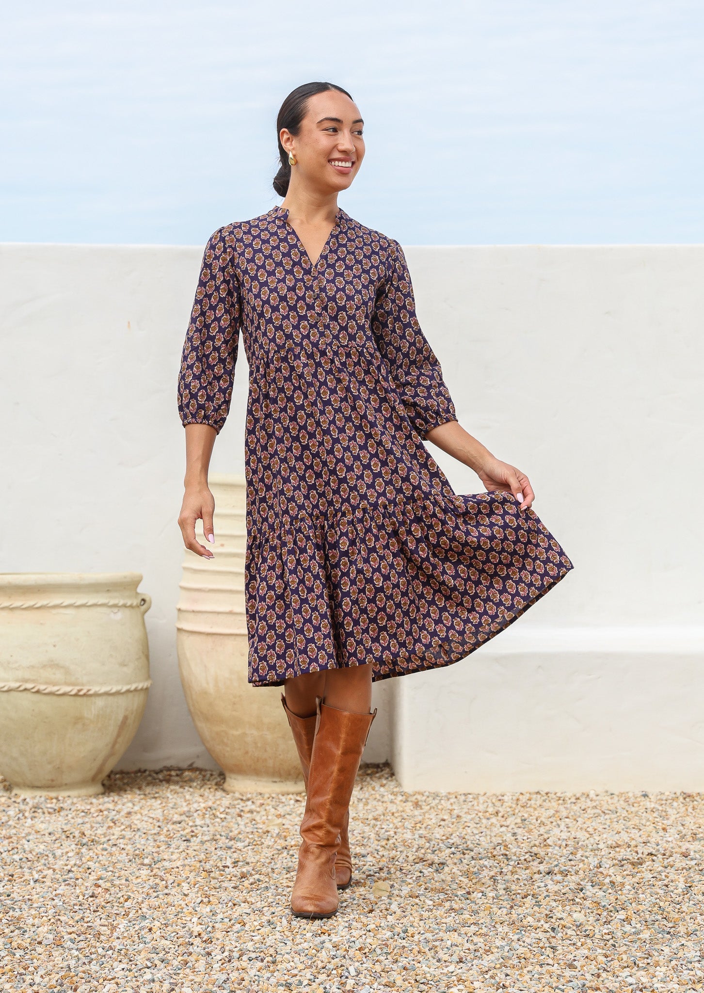 Woman wears cotton tiered over the knee length dress with 3/4 sleeves in a floral motif on a dark purple base
