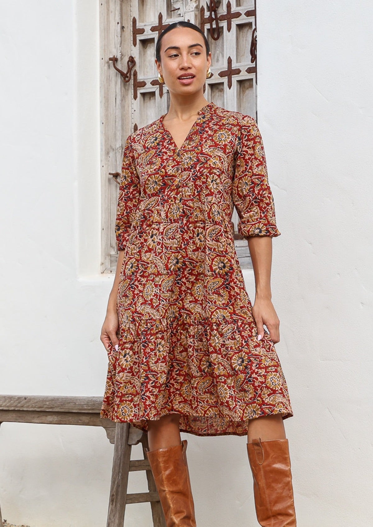 Woman wears cotton tiered dress in bright orangey red based floral print, with 3/4 sleeves, buttoned bodice and V-neckline