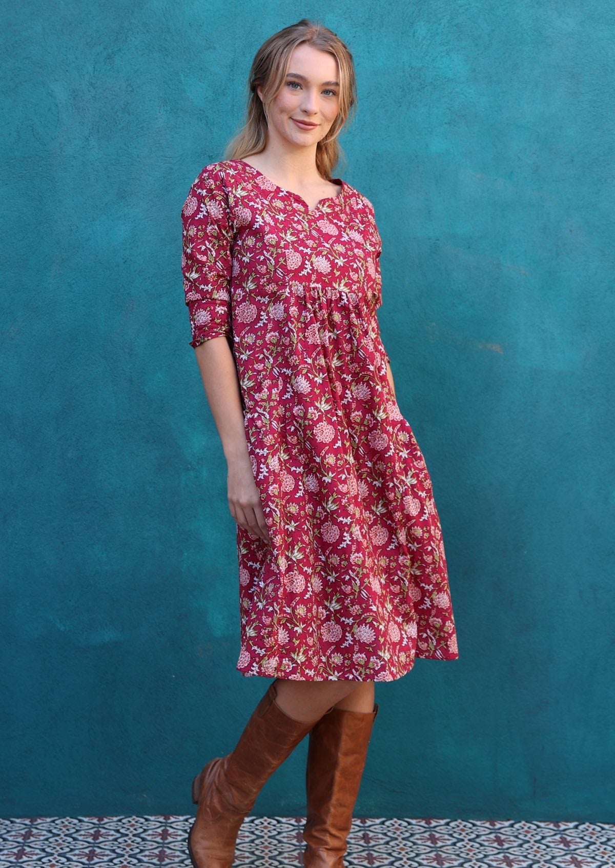 Model standing in front of blue wall wearing a beautiful red cotton dress with empire waist, 3/4 sleeves, pockets and a sweet keyhole.