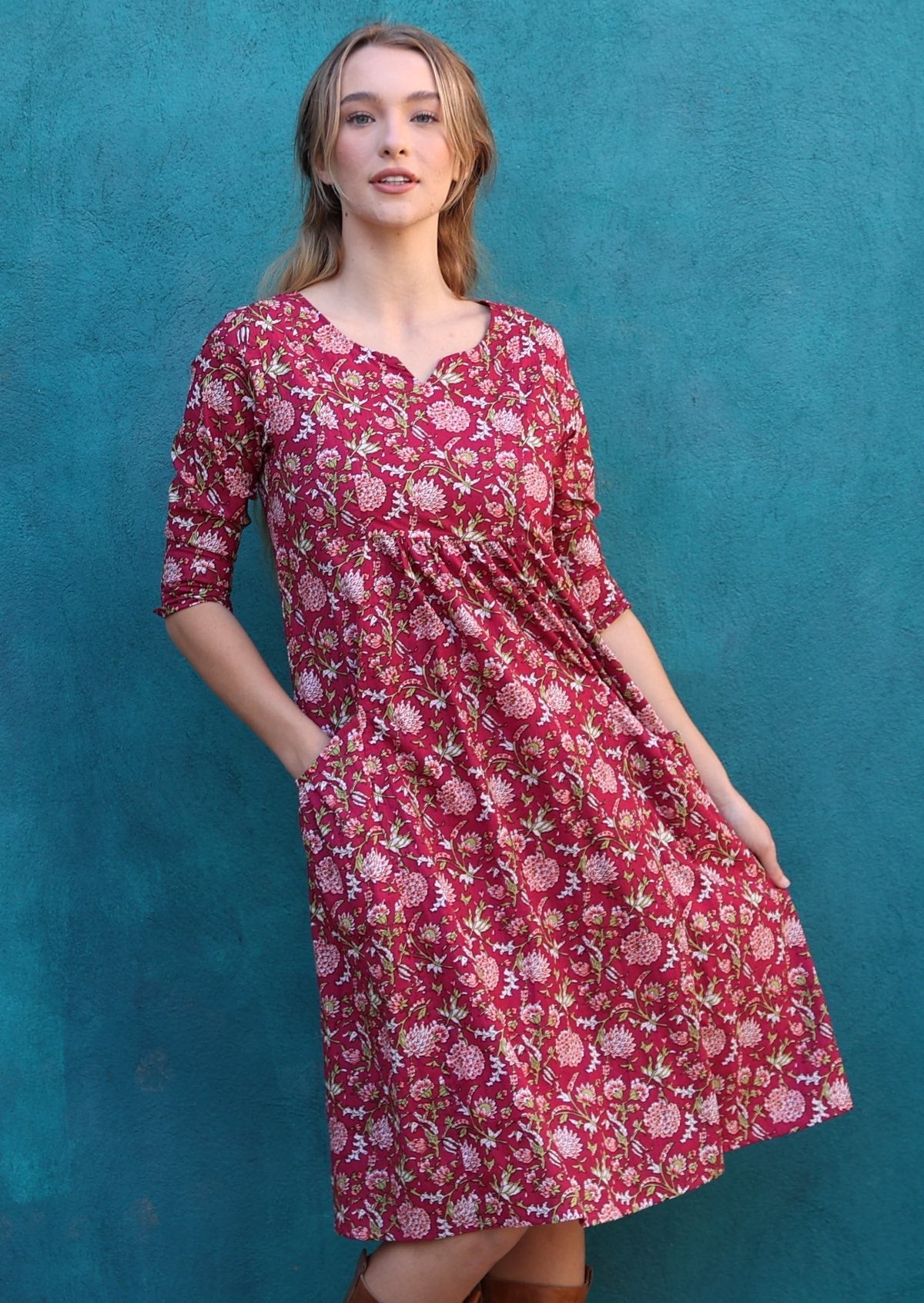 Model standing in front of blue wall wears 3/4 sleeves cotton dress with round neckline, pockets and pin tucks on waist.