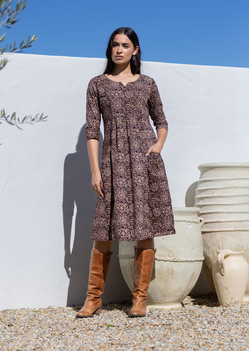 Woman wearing a brown floral cotton dress and brown boots standing against a white wall with large pots.