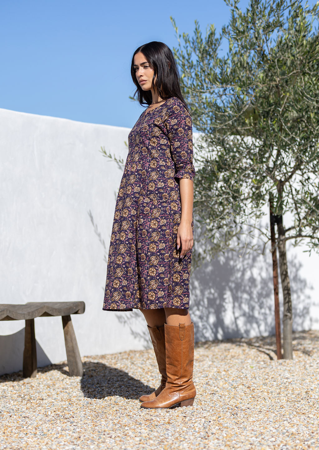 Woman in a purple cotton sundress and brown boots standing outdoors with a white wall and tree in the background.