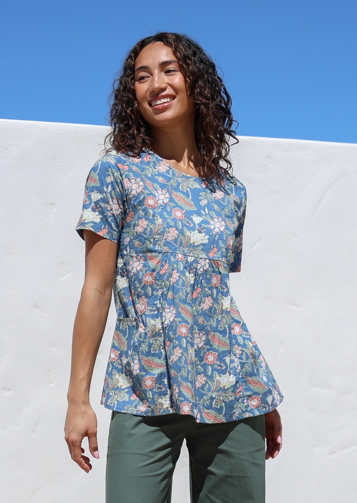Woman wearing a relaxed fit dusty blue based floral print top against a white wall with blue sky.