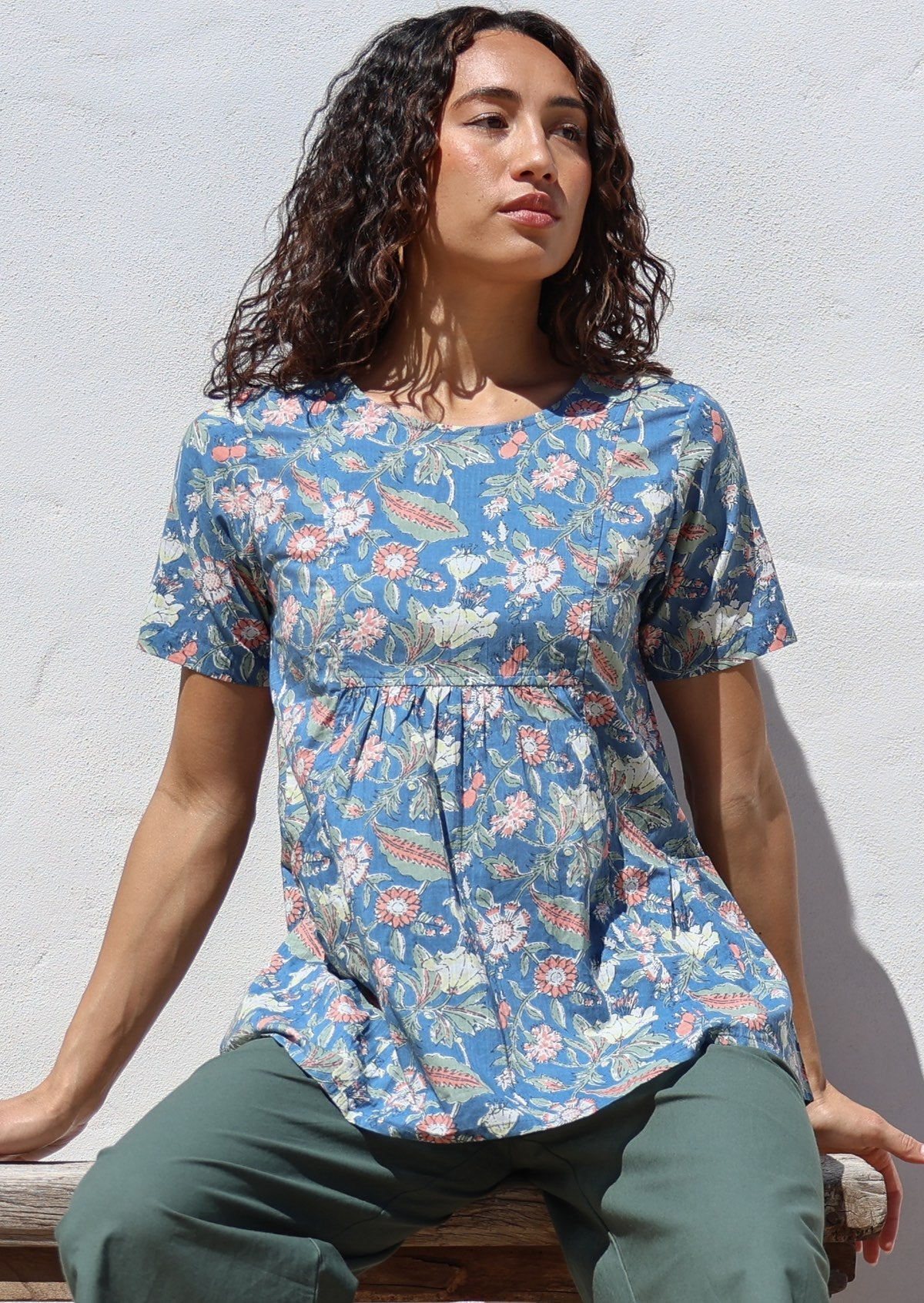 Woman sitting on a bench wearing a blue based floral print cotton top with gathers under the bust, paired with green pants