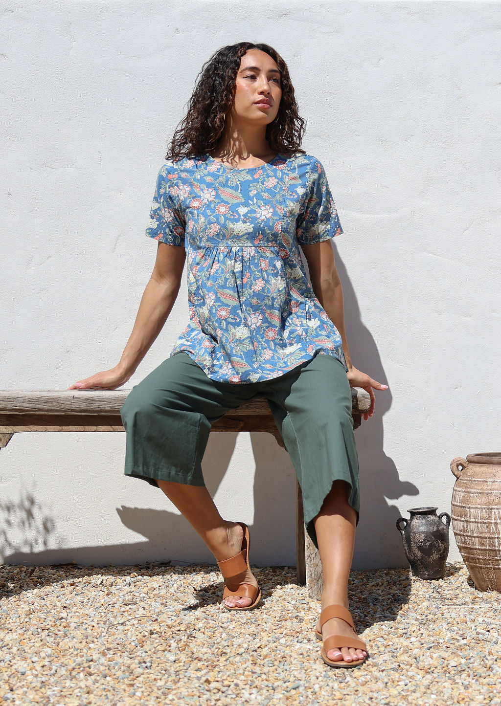 Woman sitting on a wooden bench wearing a floral print relaxed fit top and green pants against a white wall.
