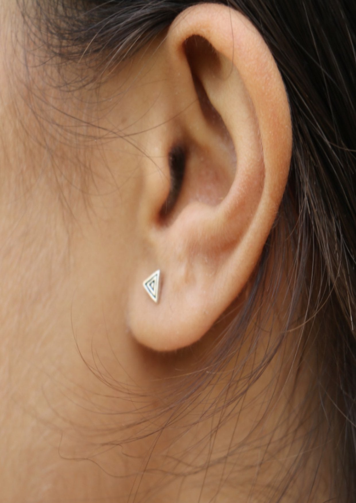 Close up of an ear wearing small sterling silver triangular studs with ripple design and butterfly backs