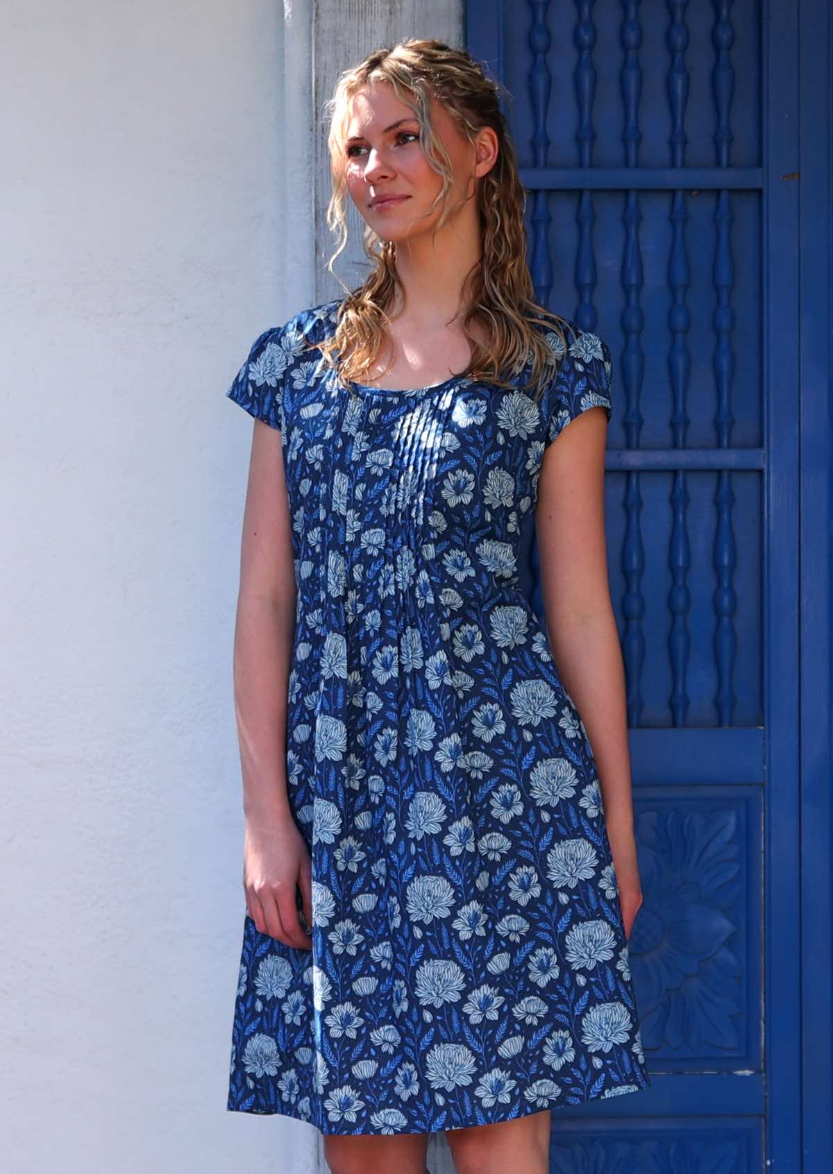 Woman with blonde hair wearing a blue cotton dress with cap sleeves in light blue and white floral design, in front of blue door