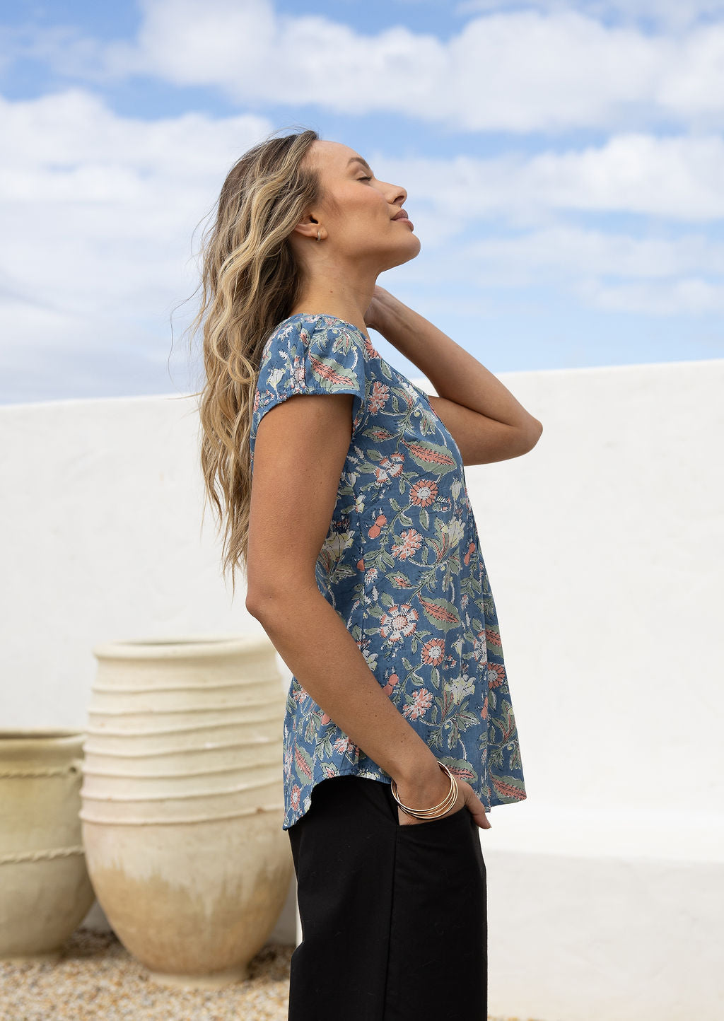 Woman standing sideways wears 100 percent cotton top with short cap sleeves and longer back hem.