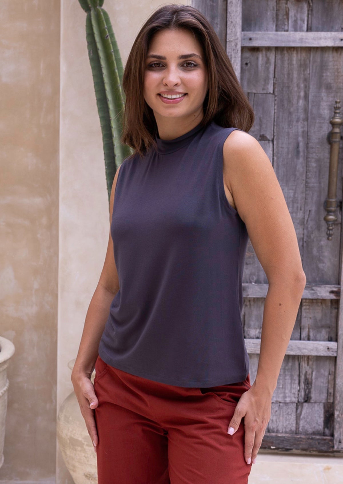 Woman wearing a sleeveless dark grey top and red cotton pants standing outdoors with a wooden door and cactus in the background.