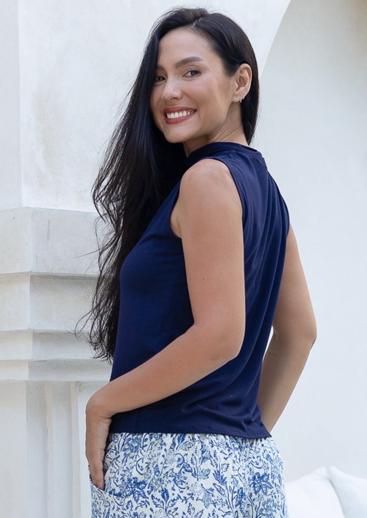 Woman wearing a navy sleeveless top with a high neckline, and floral pants standing against a white wall.