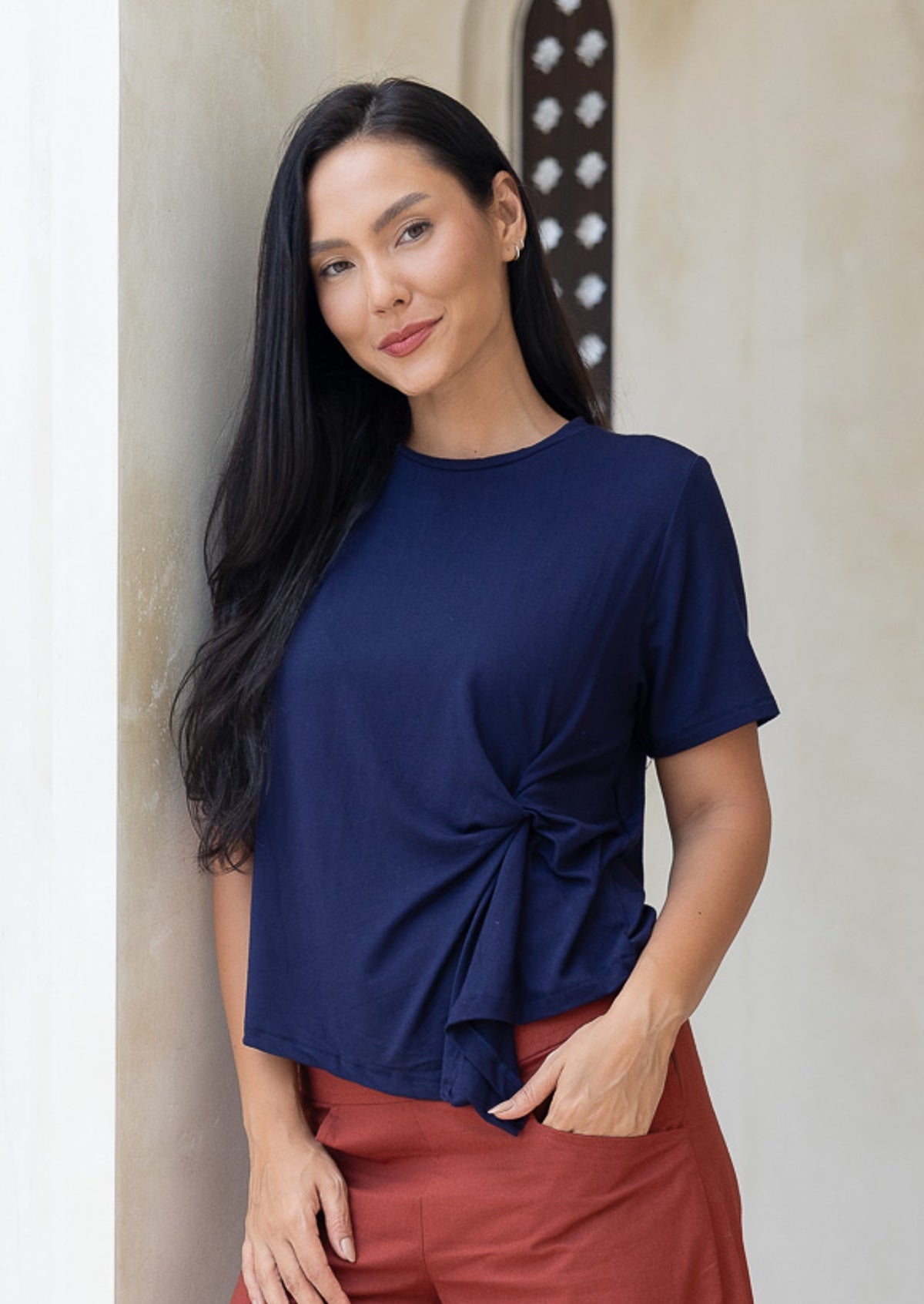 Woman wearing a navy blue short sleeve top with a knot detail in the front with hand in pocket of rust pants