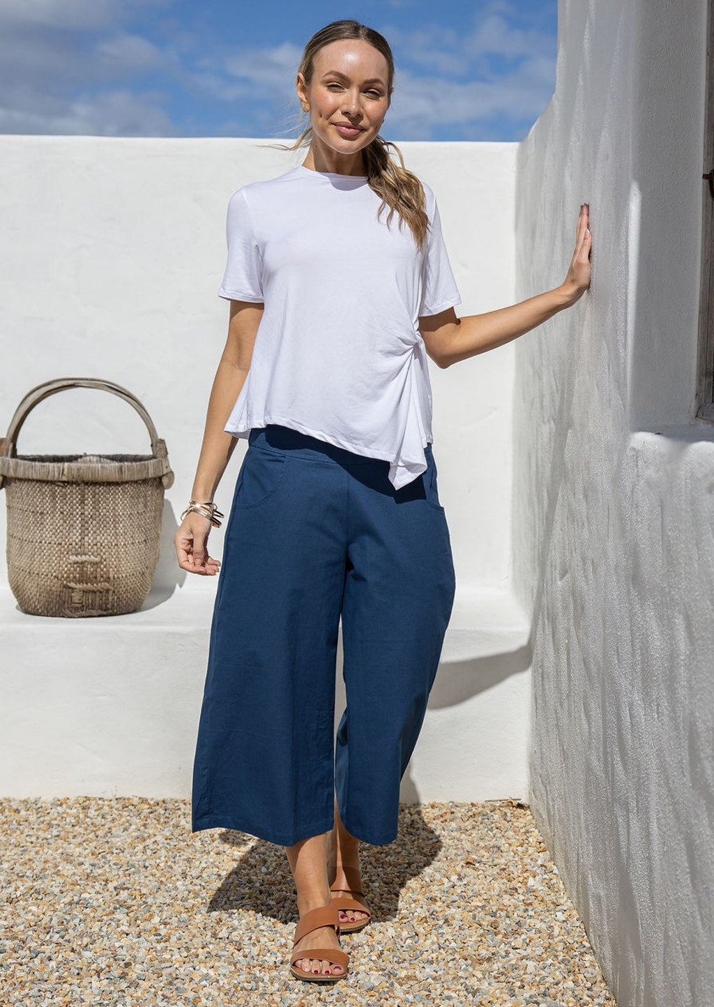 Woman in white top and blue pants standing against a white wall with a blue sky.