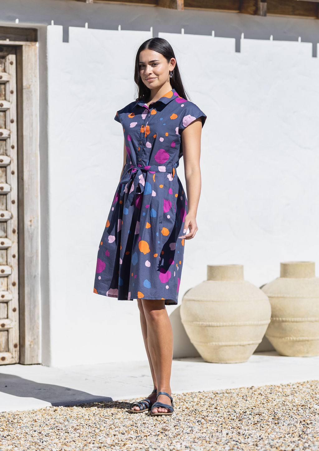 Woman wearing a plum, orange, pink and blue on a navy base spotty cotton dress standing in front of a white wall