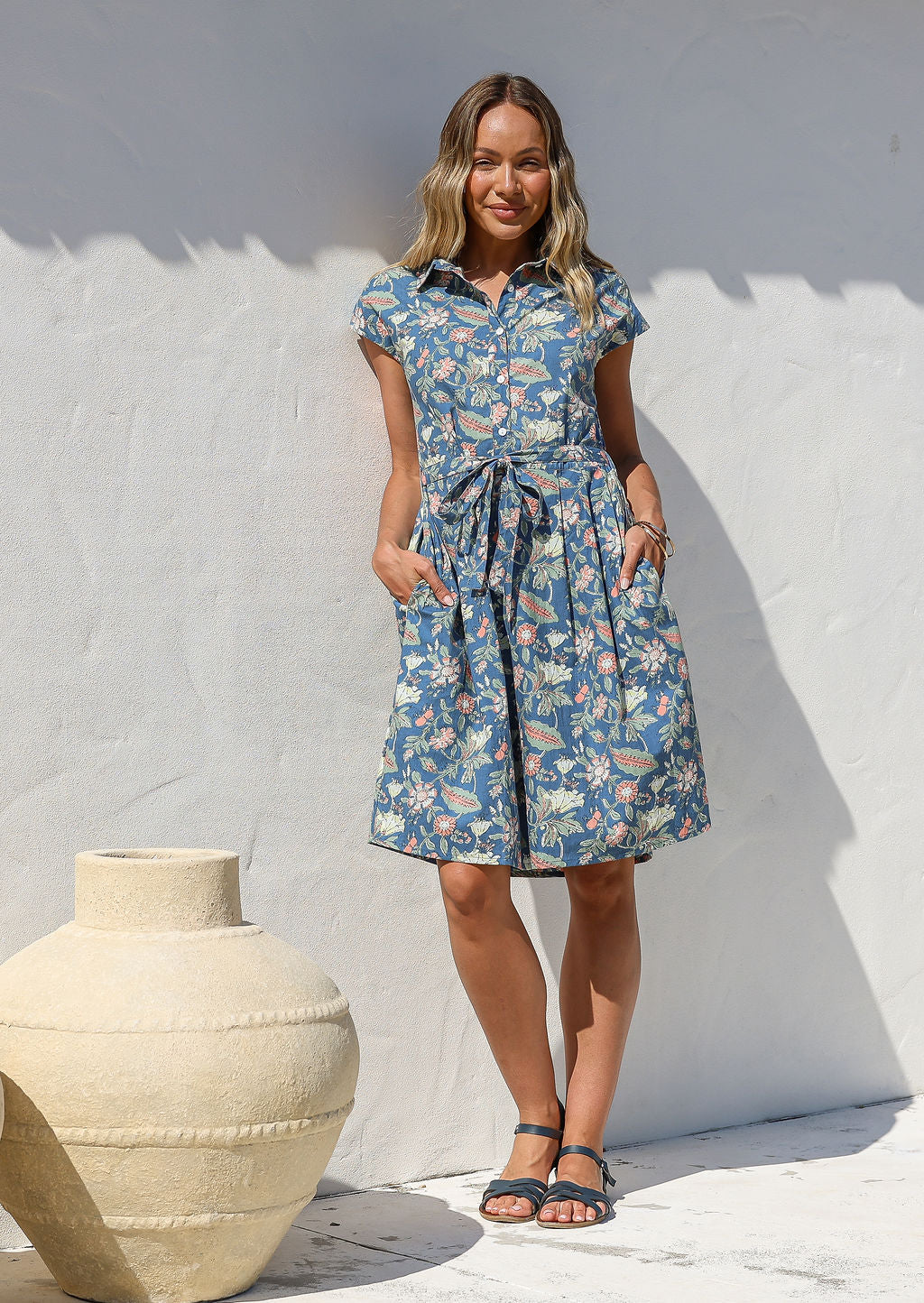 Model wears a blue, knee-length, retro style cotton dress with a buttoned bodice, pockets, cap sleeves, box pleats, and a removable waist tie.