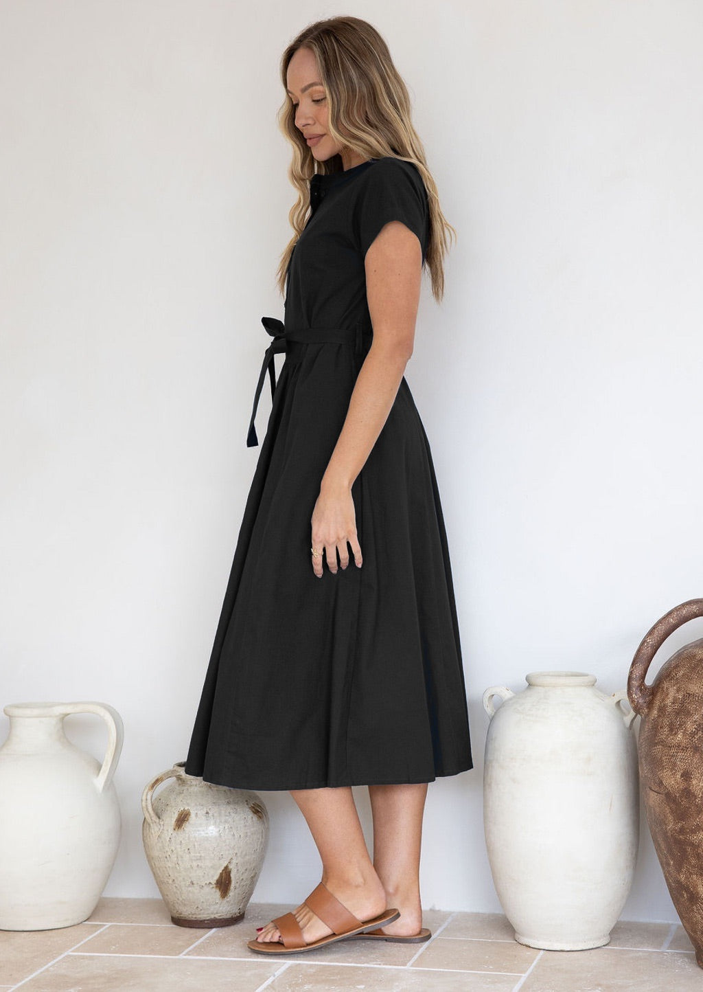 Woman standing side on, wearing a black 50's style cotton dress with cap sleeves and a waist tie