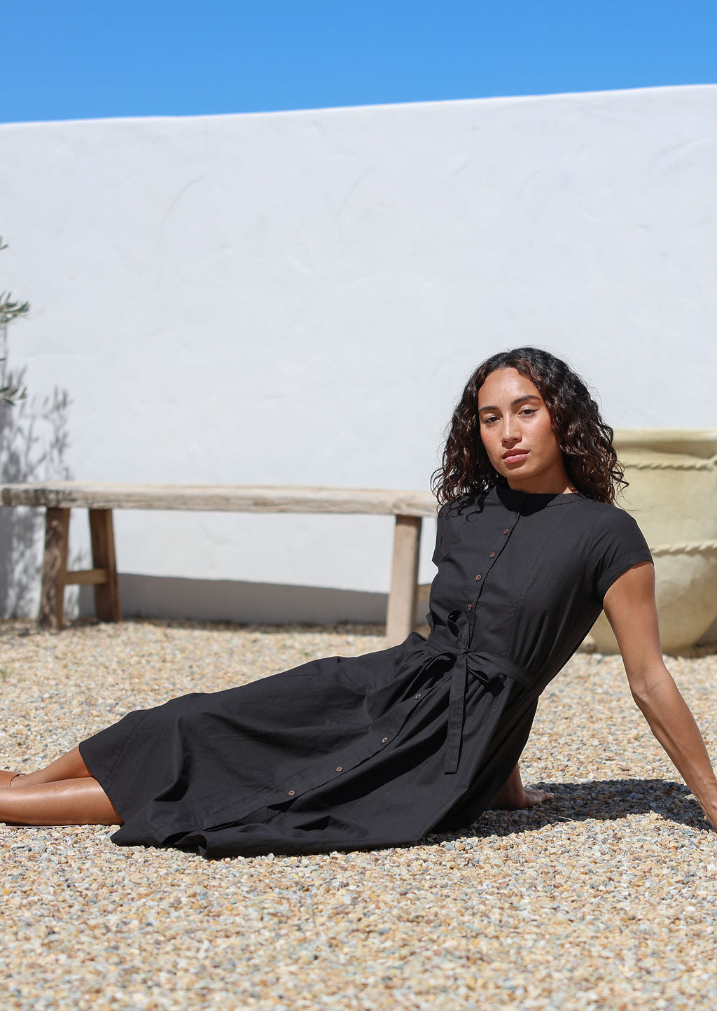 Woman in a black cotton dress sitting on pebbles with a white wall and blue sky in the background