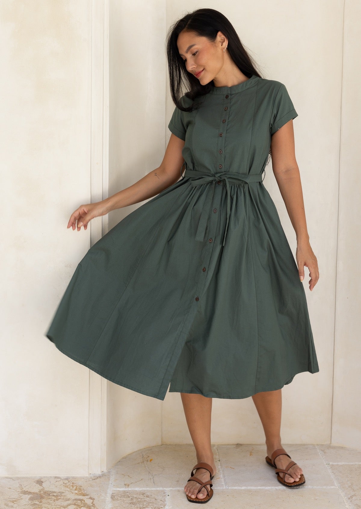 Woman wearing a moss green button through 50's style dress with a waist tie, flicking the skirt to the side