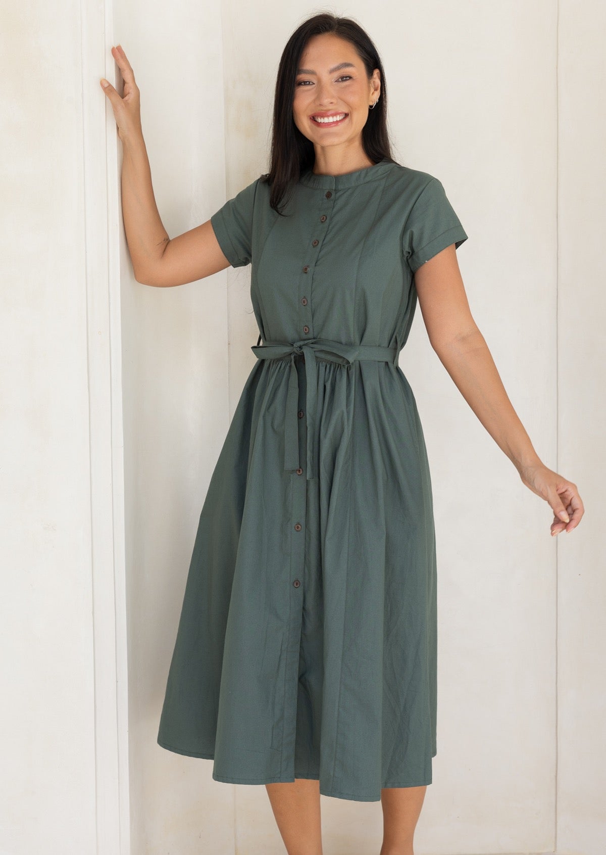 Woman wearing a moss green button through midi length dress with gathers at the waist, cinched with a waist tie