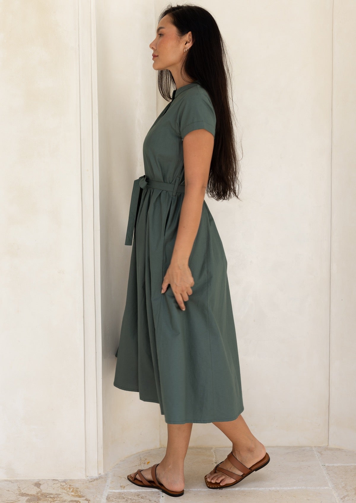 Woman standing side on wearing a green midi length dress with cap sleeves and a waist tie