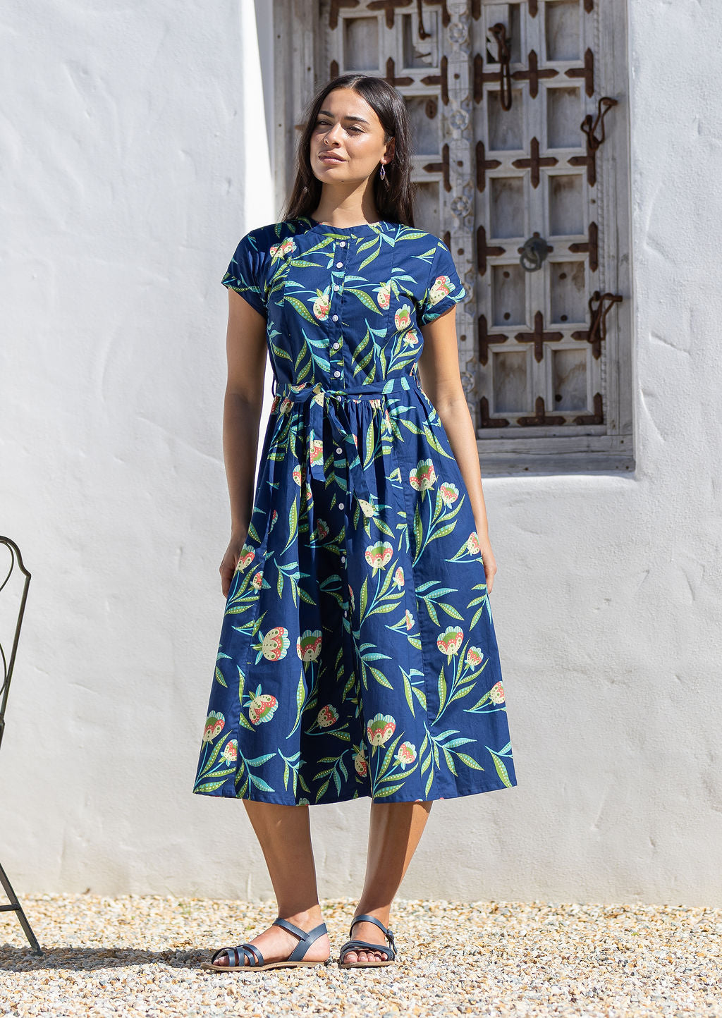 Woman wearing a blue floral cotton sundress standing in front of a white wall with a decorative metal gate.