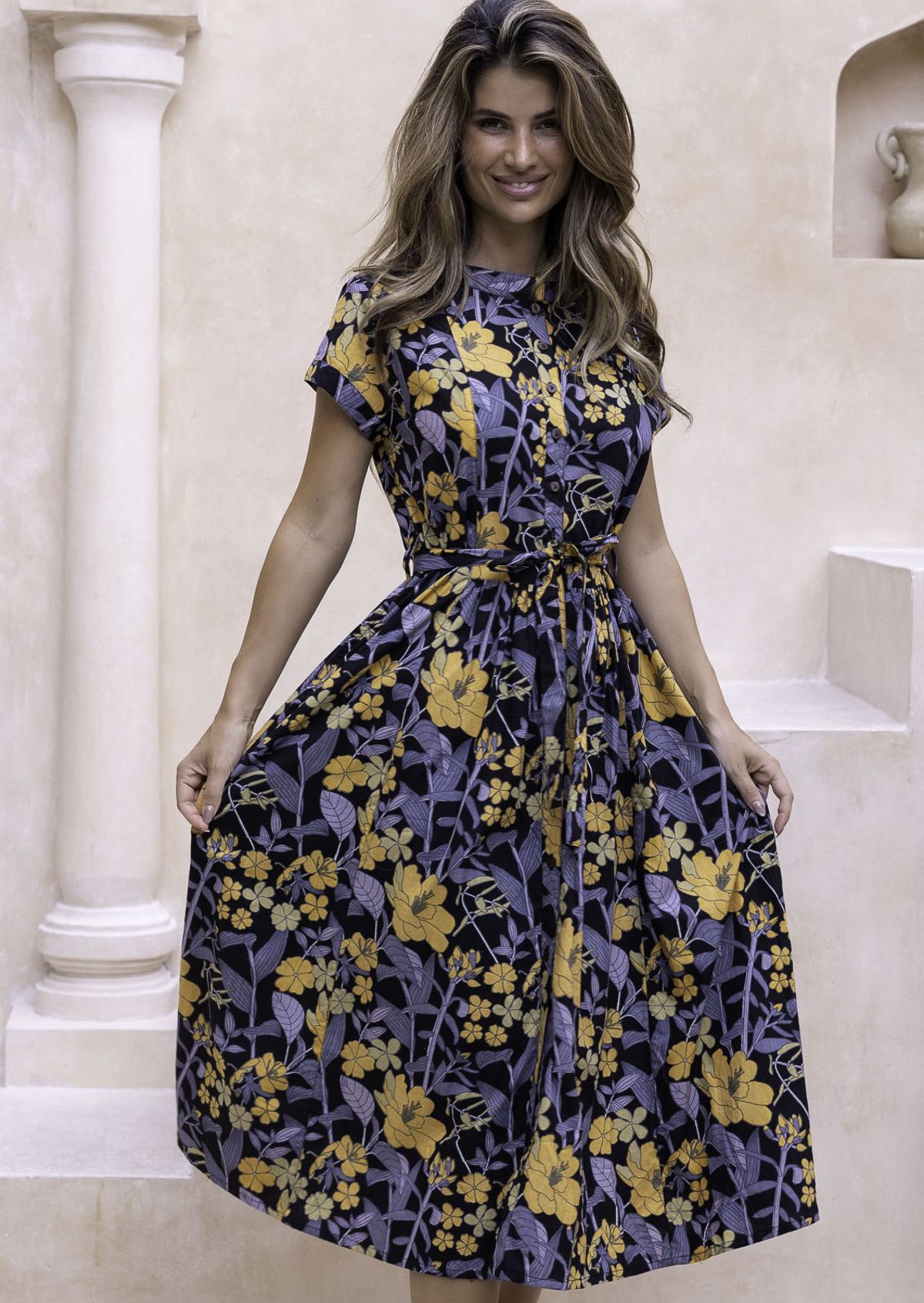 Model wears mid length cotton dress with yellow, green and grey floral print on black base.