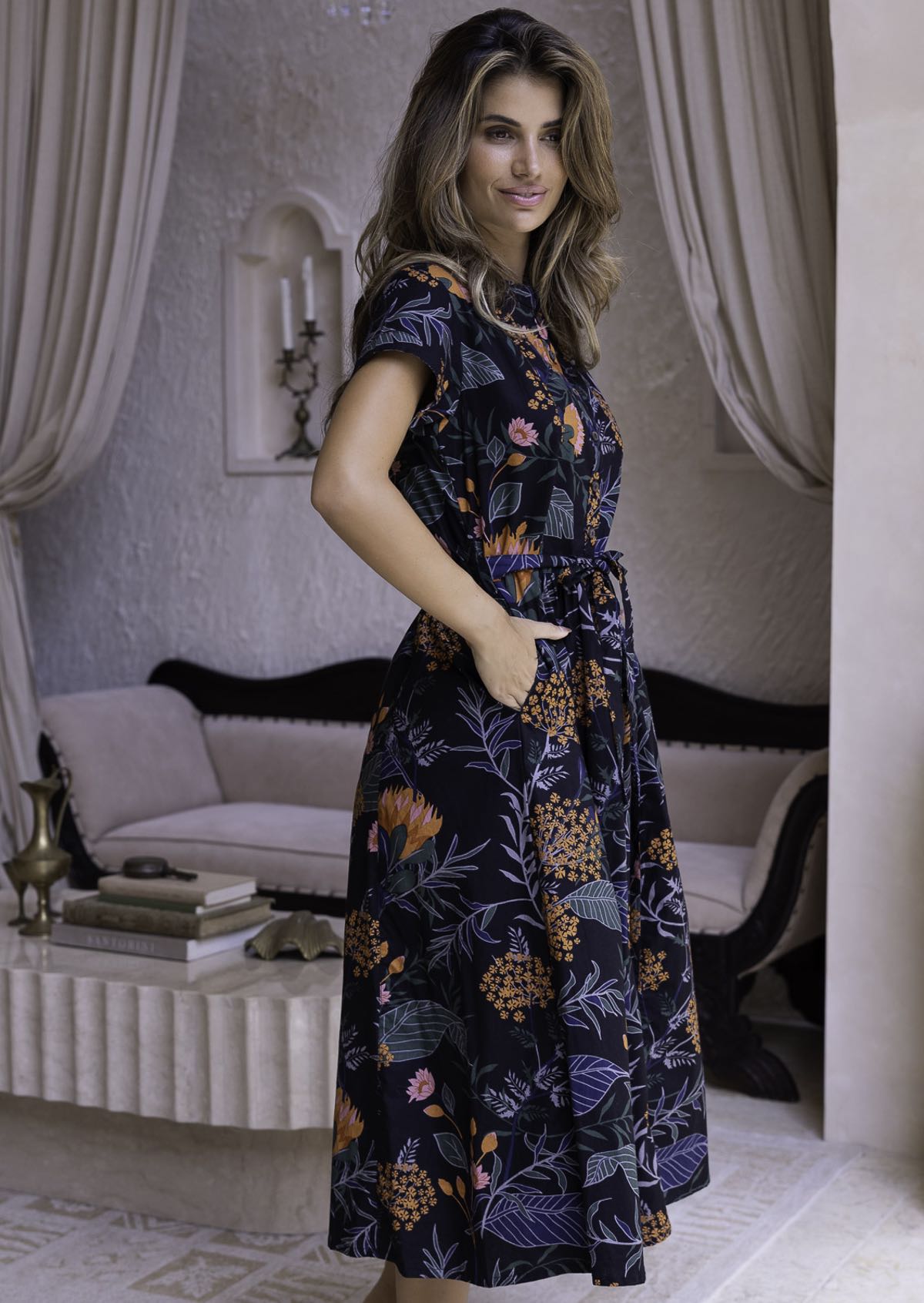 Model stands slightly side ways in 100 percent cotton retro button through dress with orange and pink florals; blue, green and grey leafy design on black base.