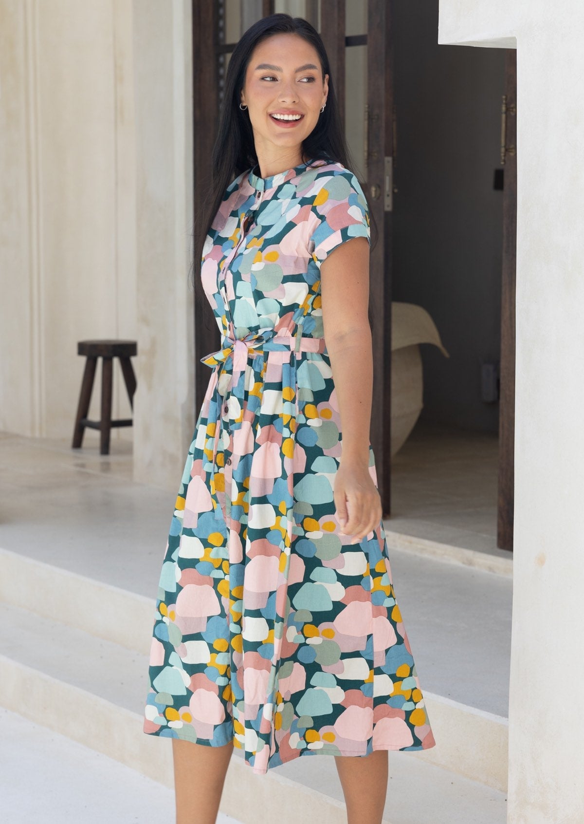 Woman wears pastel print 50's style midi length dress with cap sleeves, cinched with waist tie