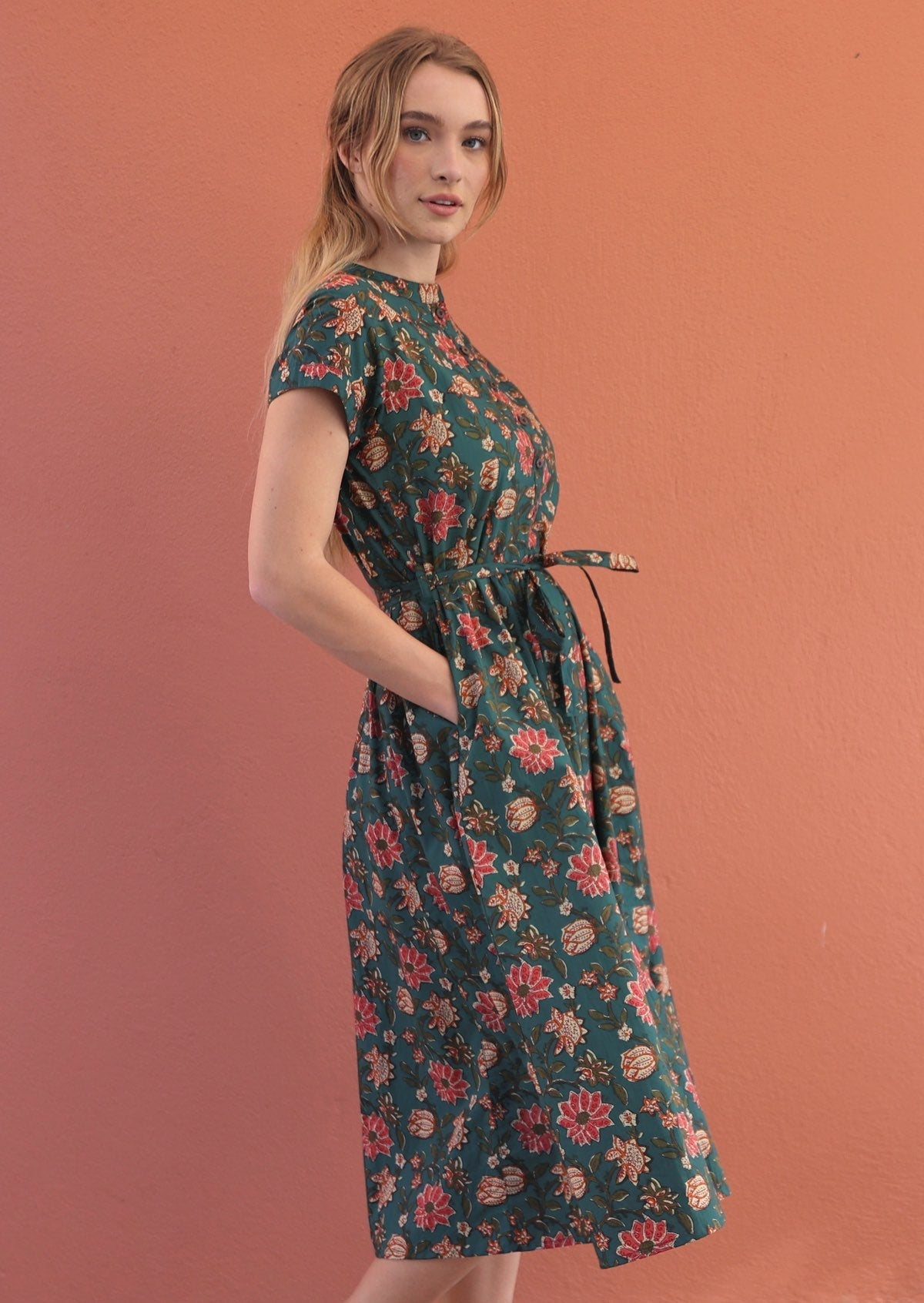Side pose of model wearing a button through cotton dress with tieable waistband and hands in pockets.