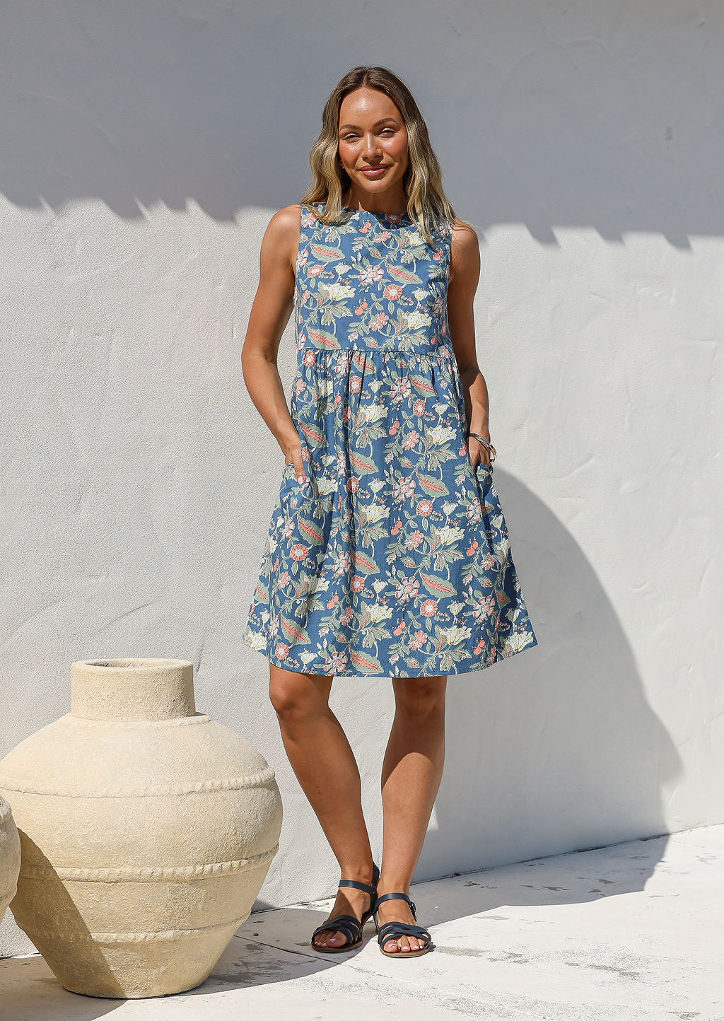 Woman in a blue floral cotton dress with sleeveless relaxed fit design and hands in pockets, standing next to a large vase against a white wall