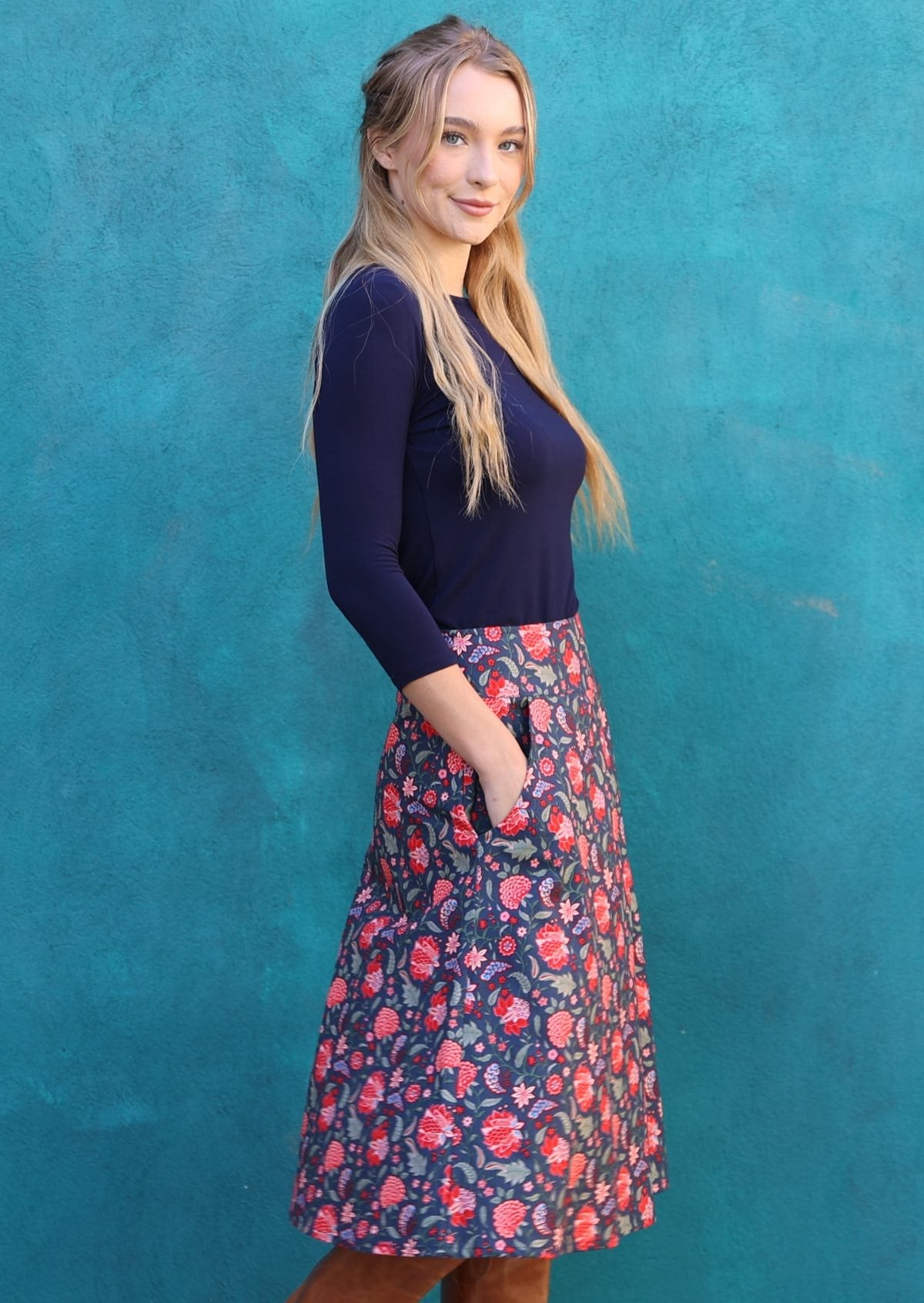Model standing sideways wears A line cotton skirt with pockets and red floral print. Pairs beautifully long sleeve navy blue top.