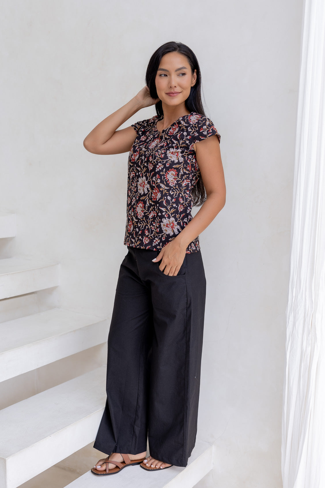 Woman posing in cap sleeved cotton top in dark brown based indian floral print, paired with wide leg black cotton pants.