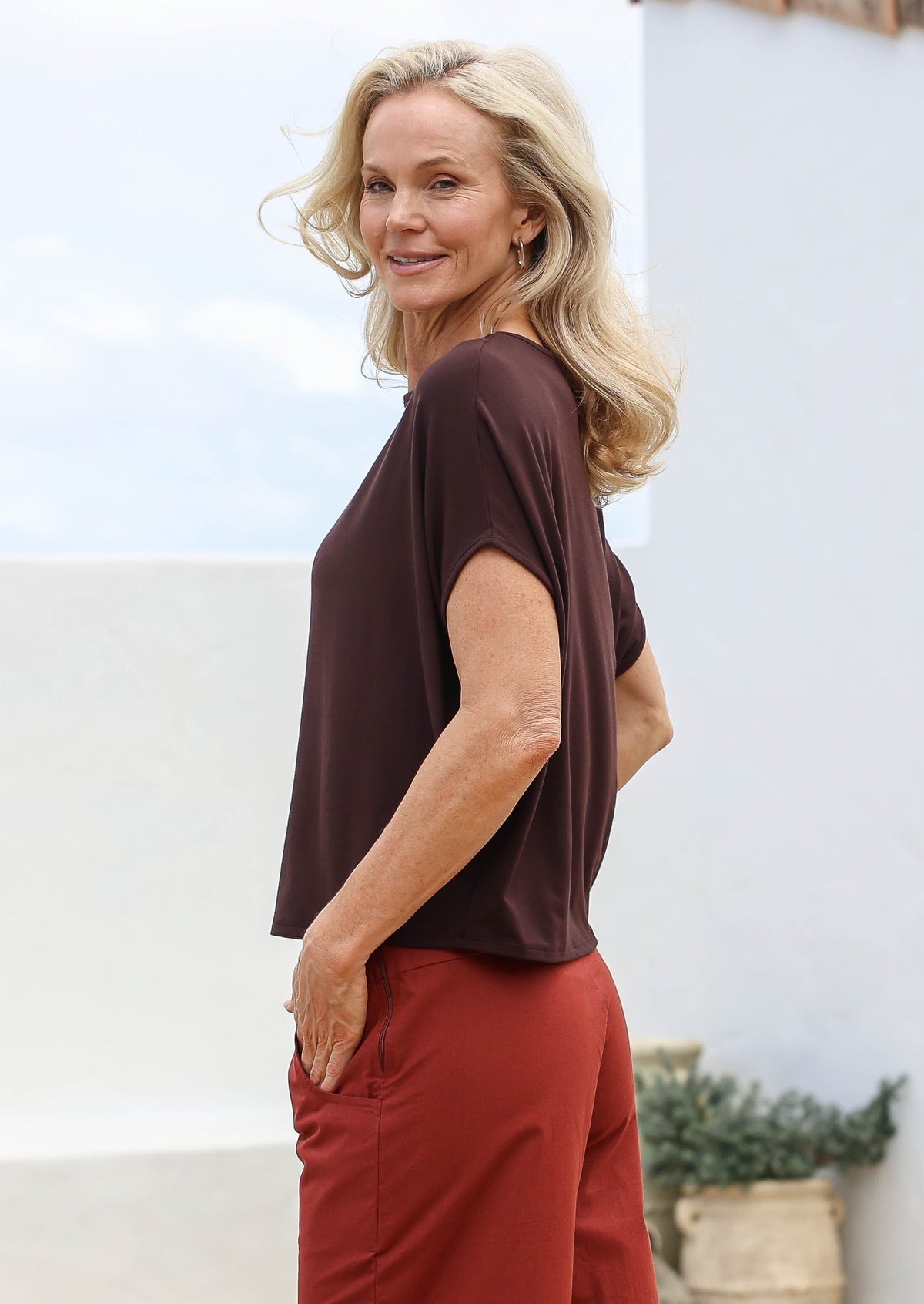 Woman wearing a brown top and burnt henna pants standing outdoors with a light background
