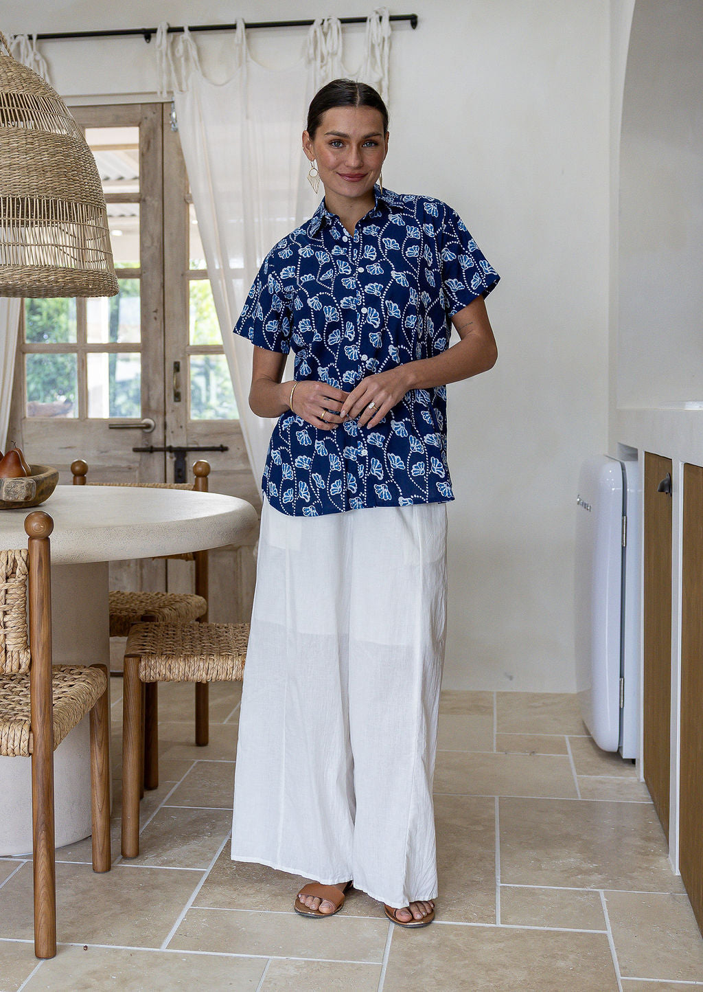 A woman stands in a bright, modern kitchen wearing a navy blue cotton short-sleeve relaxed fit button-up shirt with a white leaf pattern and white wide-leg pants.