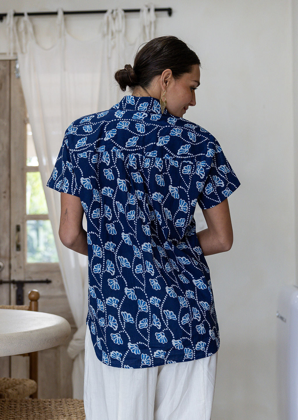 A woman viewed from behind wearing a navy blue cotton short-sleeve shirt with a white leaf block-print pattern, paired with white pants