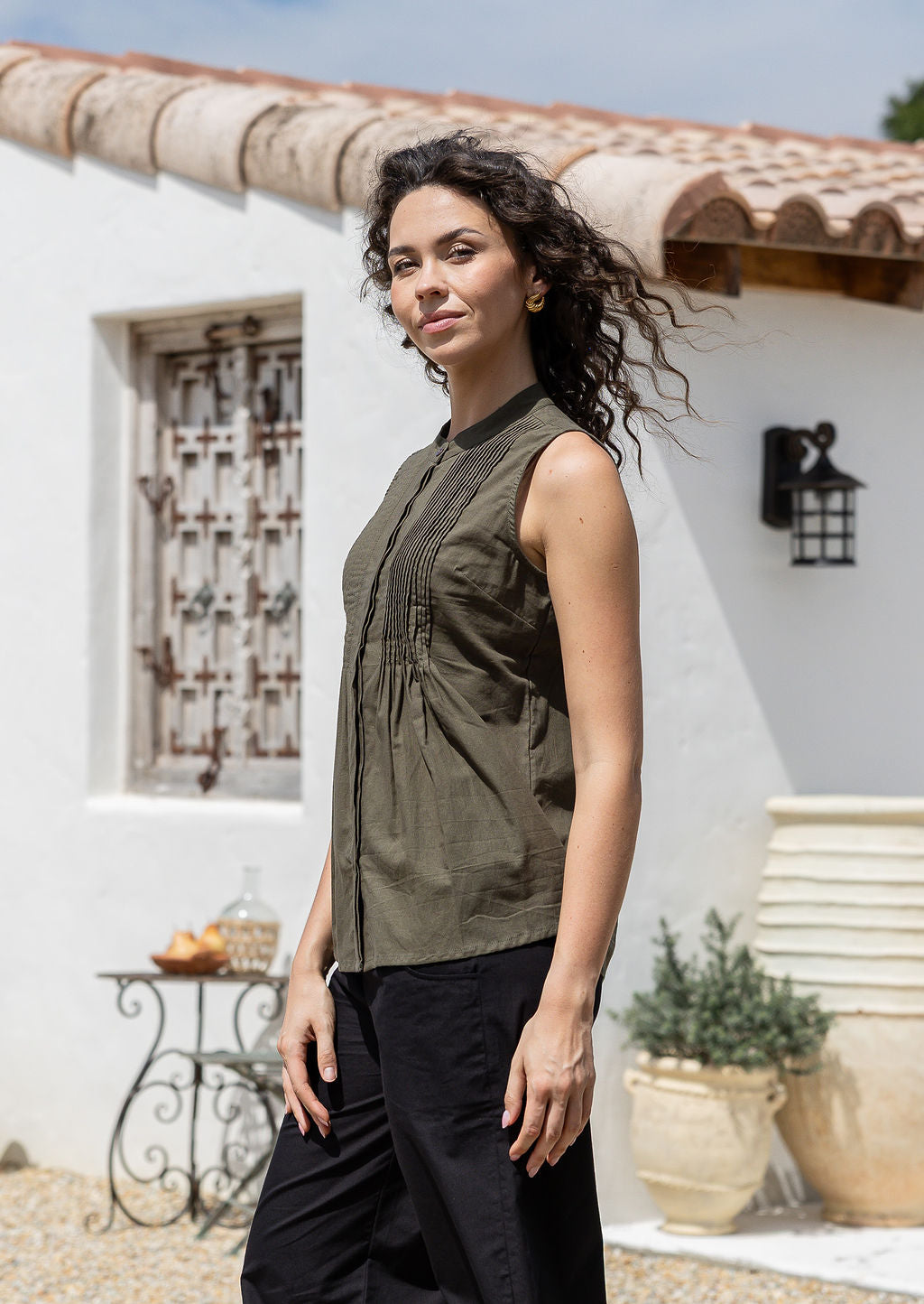 Woman stands sideways wearing 100% cotton, sleeveless top with buttoned mandarin collar, round neckline and decorative small pleated design in front.