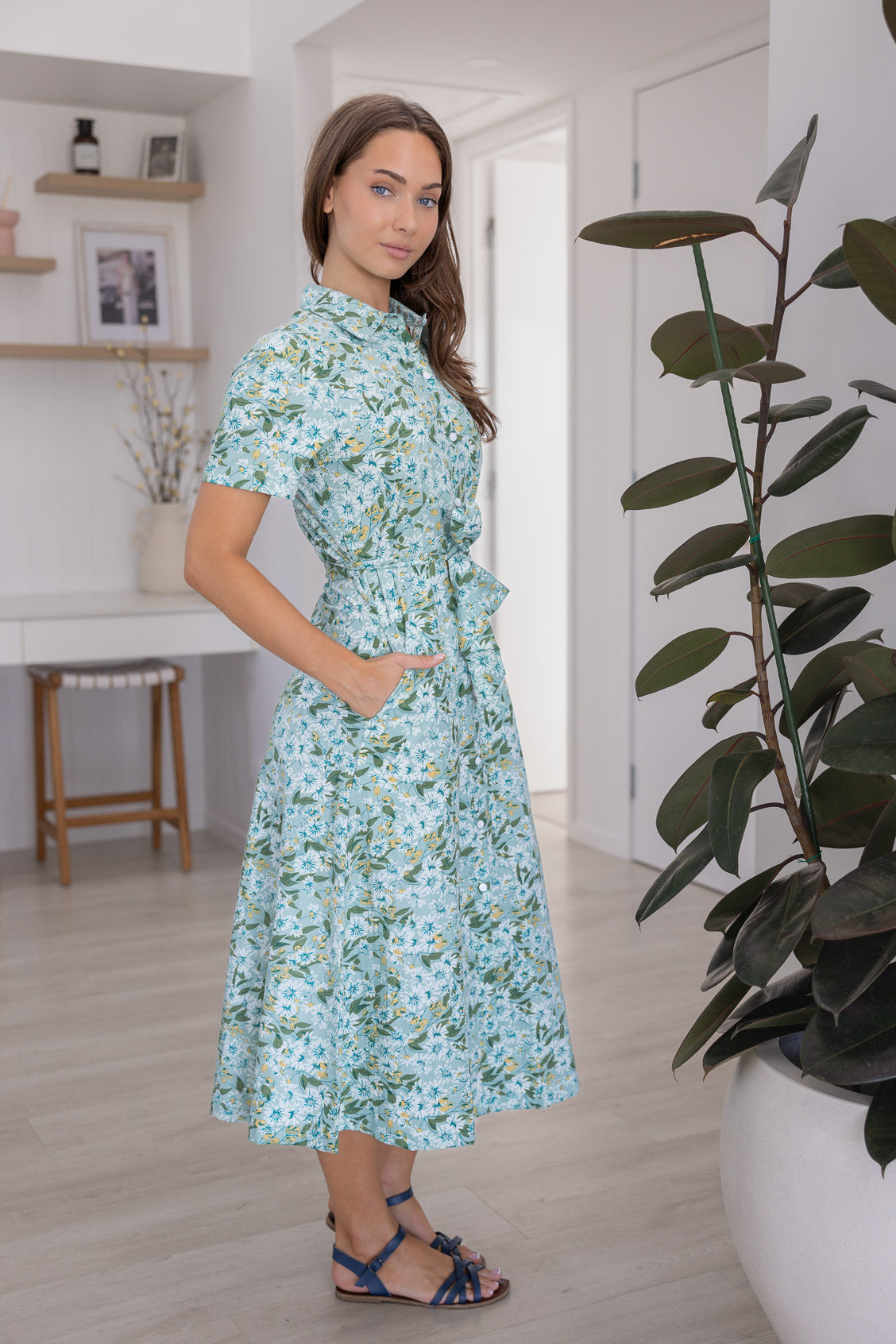 Model stands sideways wears 1950s inspired, button through shirt style dress with collar, short sleeves, waist tie and pockets.