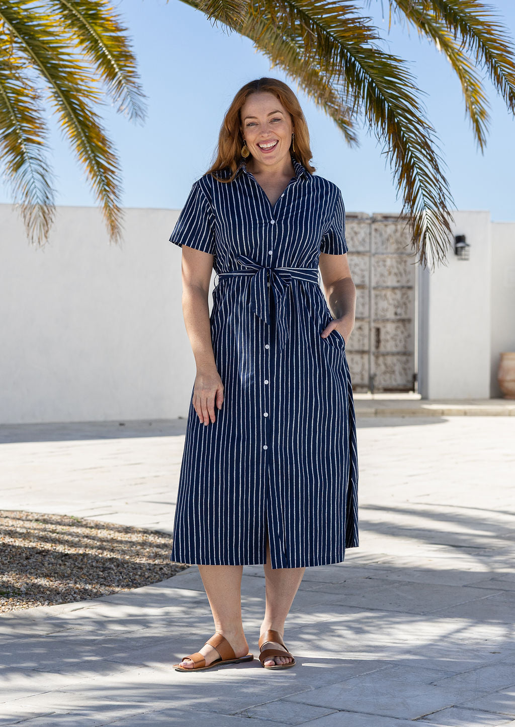 Woman wearing 100 percent cotton button through shirt dress with collar and short sleeves, in a navy based white striped print.