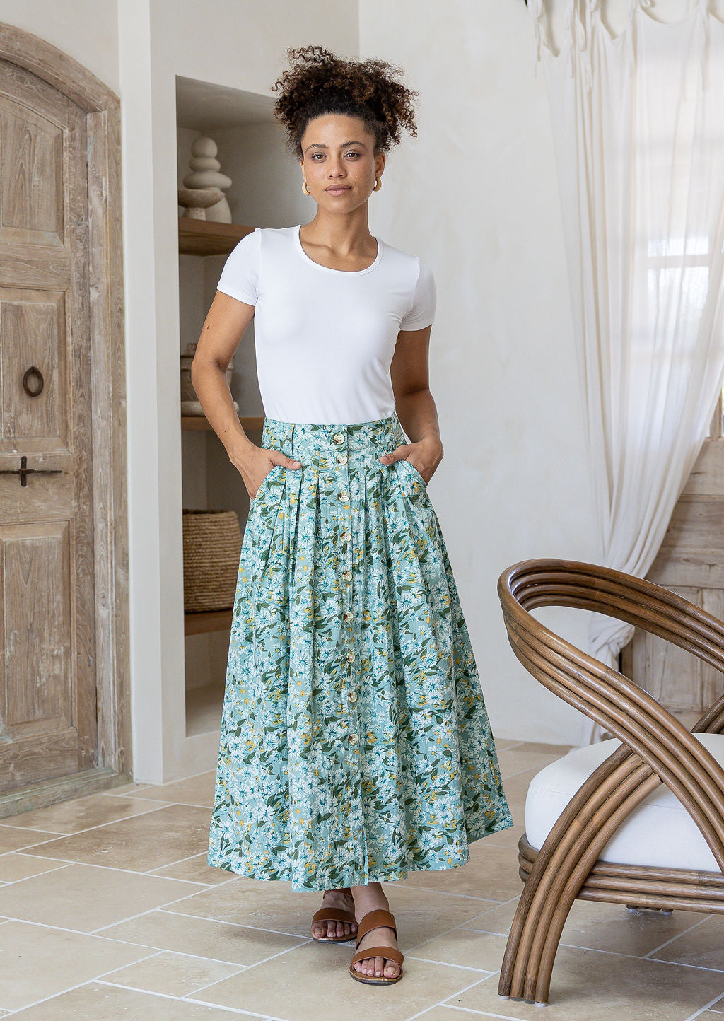 Woman standing with both hand in pockets in a minimalistic room wears 100% cotton button through skirt in sage green based floral print.