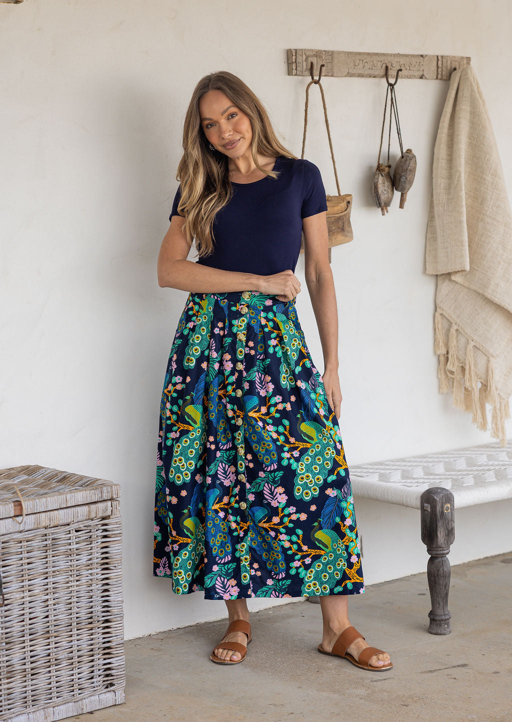 Woman standing wears 100% cotton button through skirt in navy based floral peacock print.