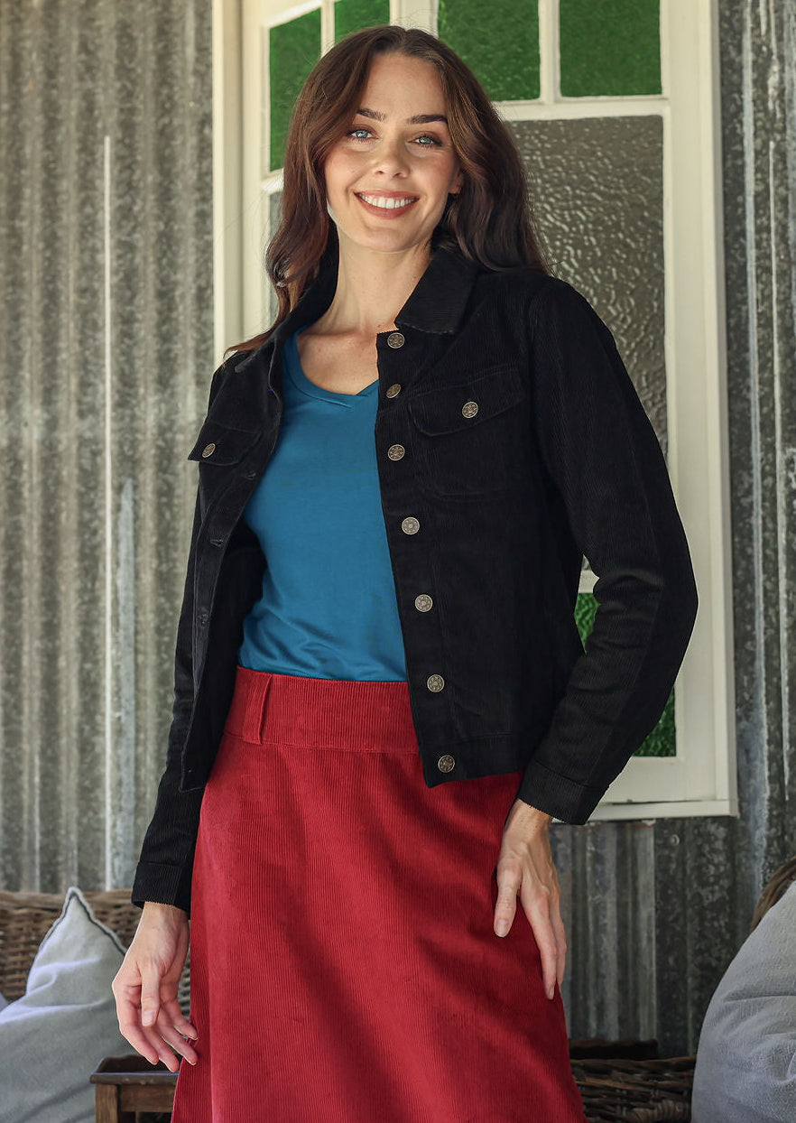Woman wearing a black cotton corduroy jacket, blue top, and red skirt standing in a room with a textured wall and window.