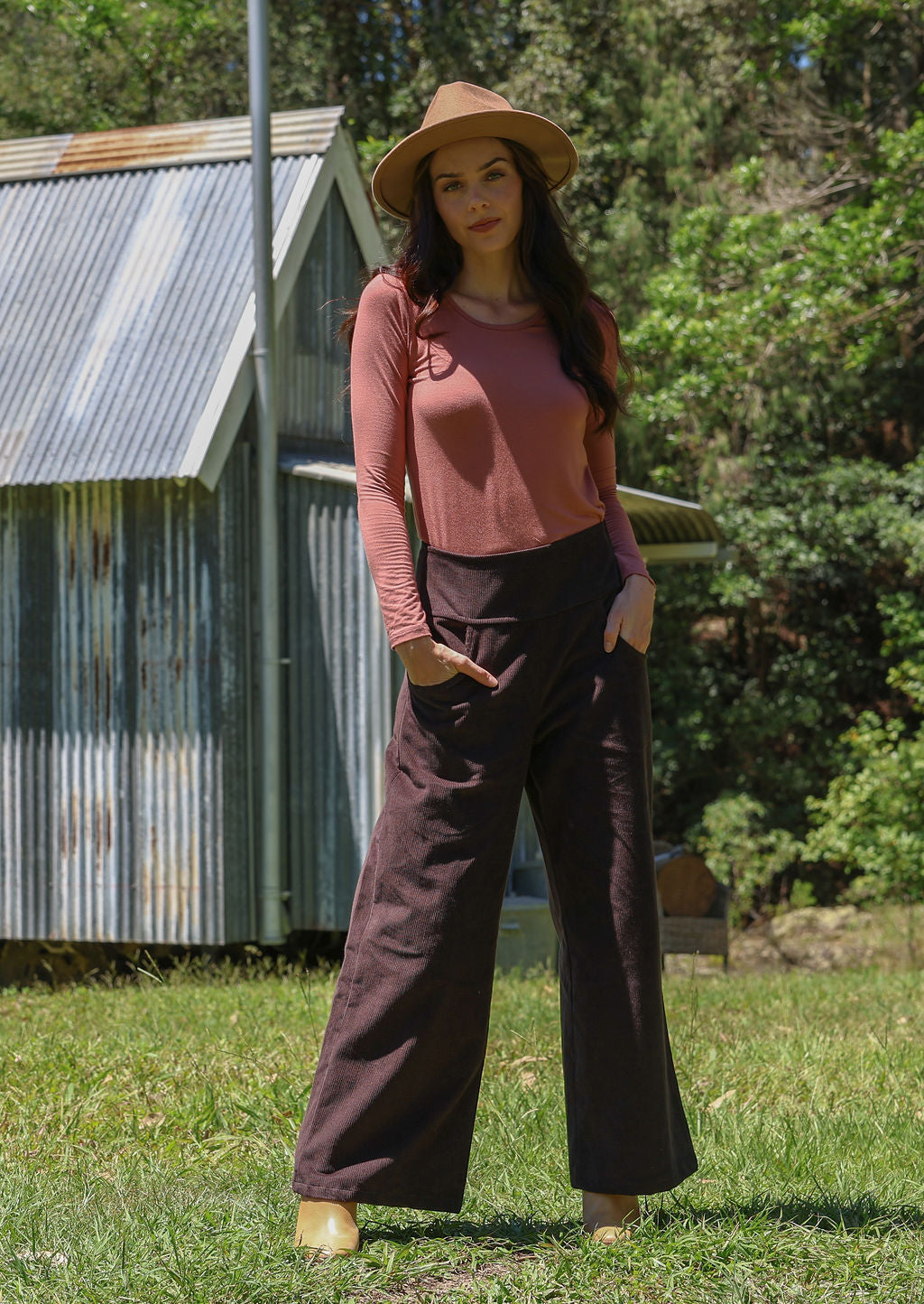 Woman in a pink top and brown pants standing outdoors with a rustic building in the background